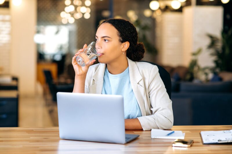 Healthy brazilian or hispanic business lady, sits at a workplace in the office, drinking a glass of fresh clean water, keeping balance of the daily water. Health care, daily water intake in the body business woman drinking glass of water at work