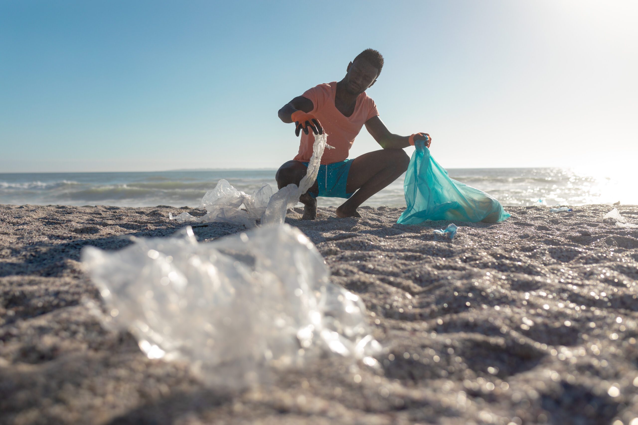 African american man crouching while collecting garbage in plastic bag at beach on sunny day. unaltered, responsibility and environmental issues concept. African american man crouching while collecting garbage in plastic bag